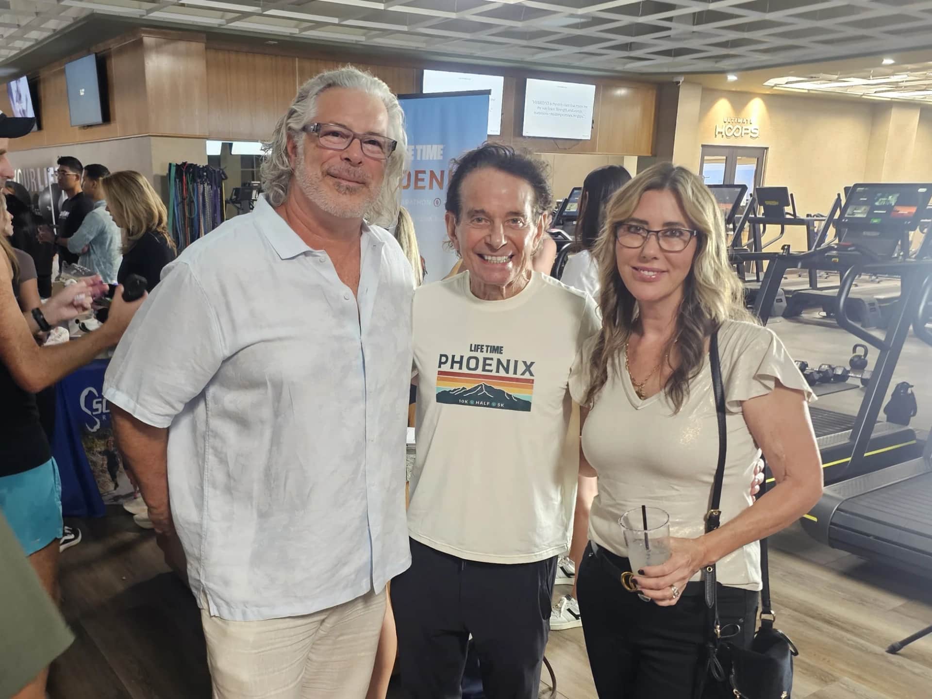 Three attendees pose at the Life Time Phoenix 10K kickoff inside a fitness center, with event signage and cardio equipment behind them.