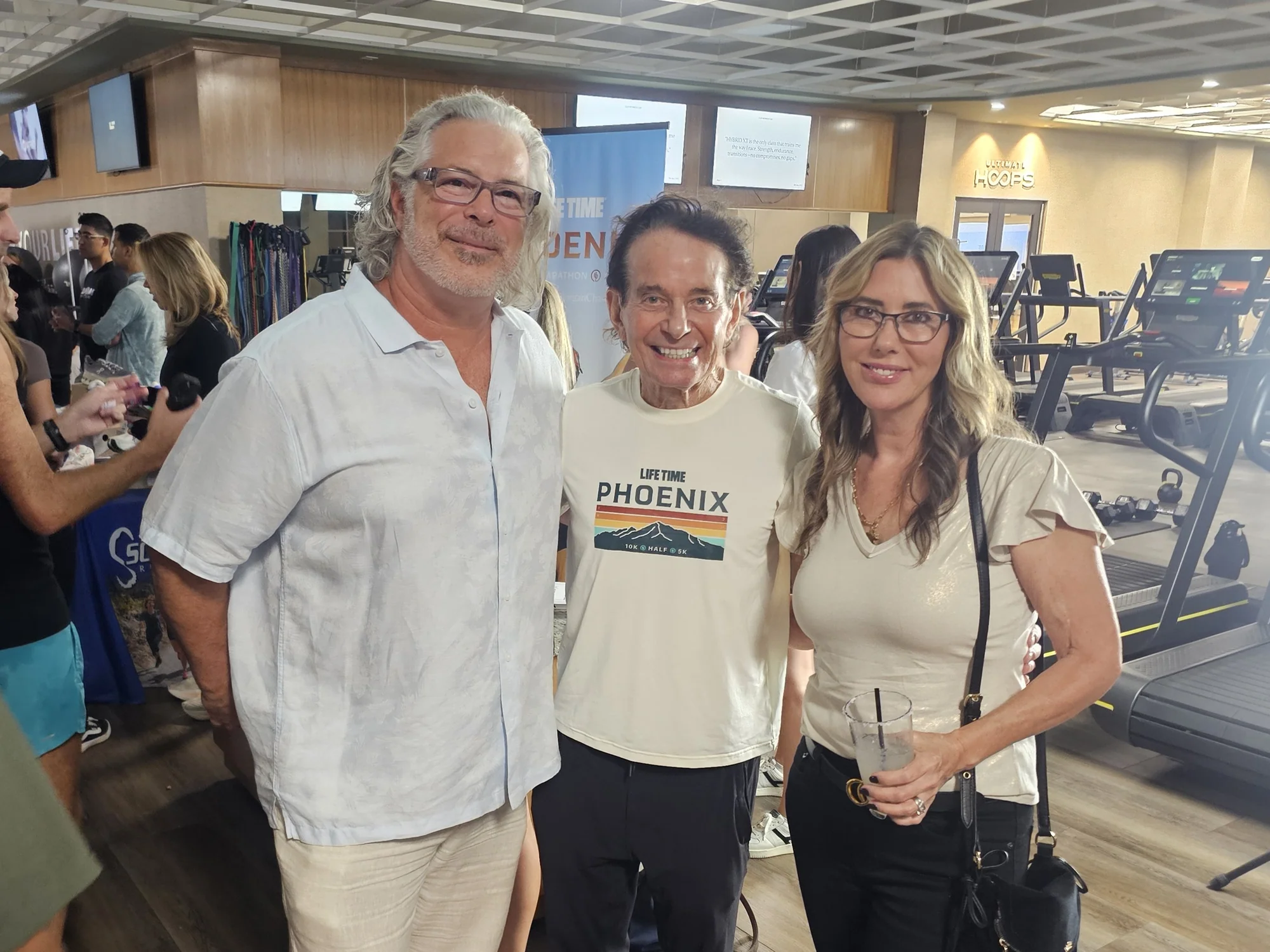 Three attendees pose at the Life Time Phoenix 10K kickoff inside a fitness center, with event signage and cardio equipment behind them.